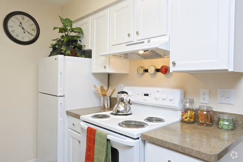 A clean kitchen with white cabinets, a white refrigerator, and a white electric stove with a kettle on it. There are three glass jars with dried pasta and vegetables on the countertop, a container holding wooden utensils, a wall clock, and a green and red towel hanging on the oven door.