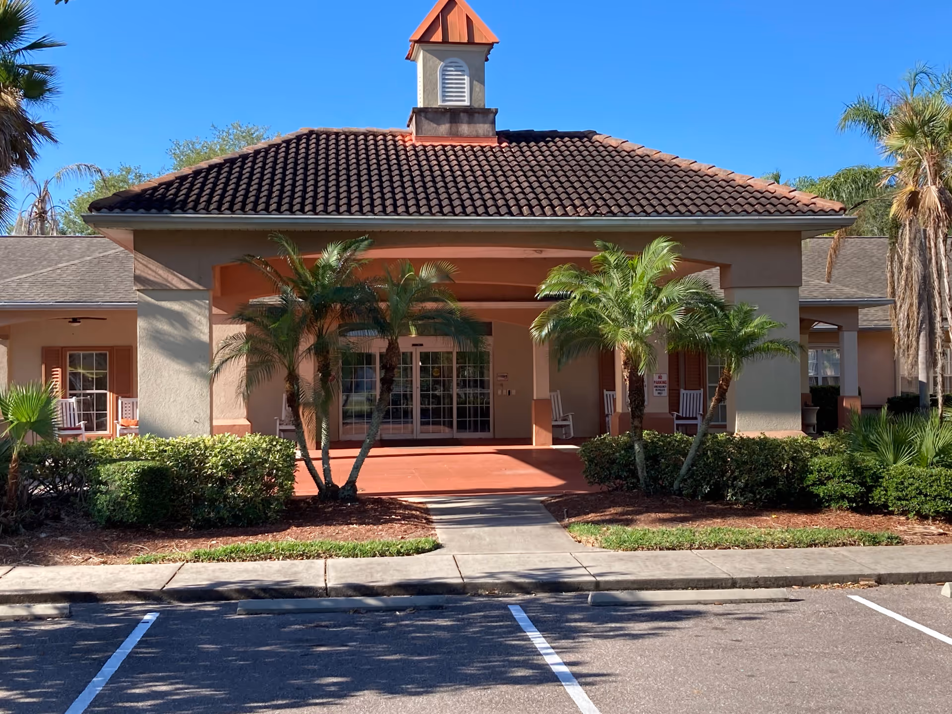 Front exterior view of Hawthorne Estates of Brandon (Independent Living) showing a covered entrance with a tiled roof, palm trees, shrubs, and a sidewalk leading to glass double doors. There are rocking chairs on the porch area under the covered entrance.