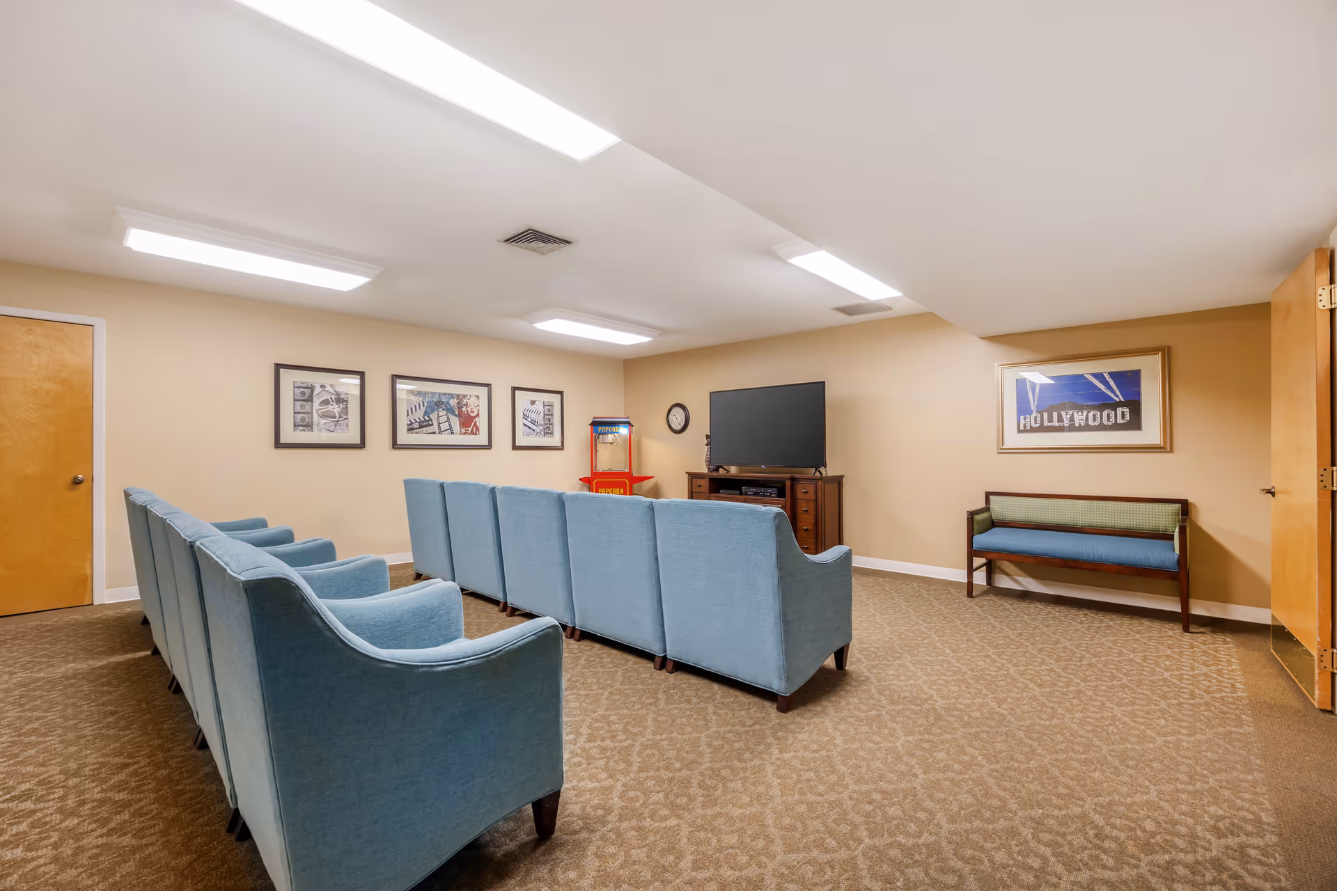 A small home theater room with two rows of blue upholstered chairs facing a flat-screen TV on a wooden stand. There is a red popcorn machine in the corner, a wall clock, and framed movie-themed artwork on the beige walls. A wooden bench with a blue cushion is placed against the right wall near an open door.