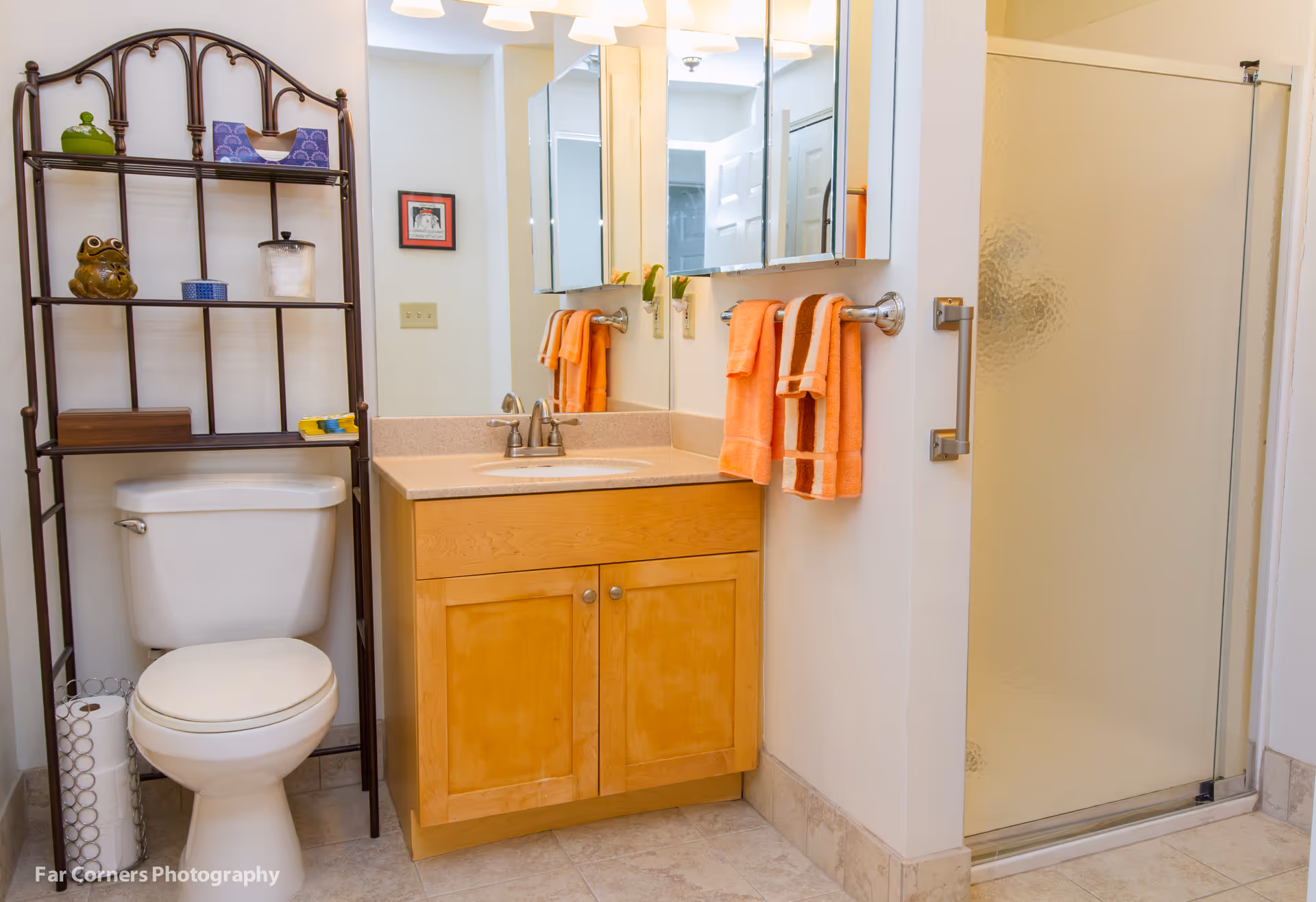 A clean bathroom with a white toilet, a wooden vanity with a sink, a large mirror above the sink, and a glass shower door. There is a metal shelving unit above the toilet holding various decorative items and toiletries. Two orange towels hang on a towel rack next to the sink.