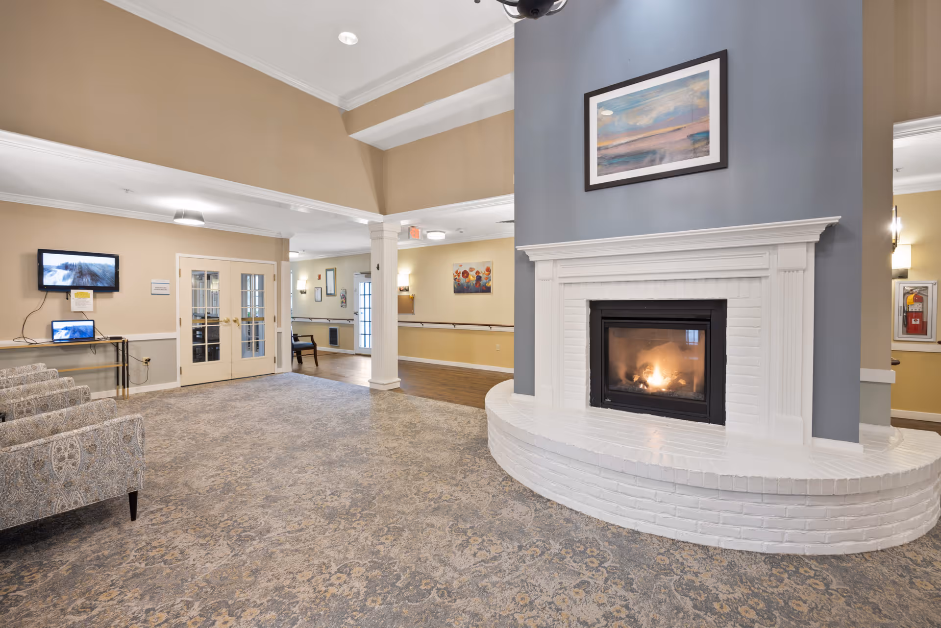 A spacious and well-lit common area featuring a white brick fireplace with a fire burning inside. Above the fireplace is a framed painting. The room has beige walls with white trim and a patterned carpet. To the left, there are patterned armchairs and a small table with a laptop and a wall-mounted TV above it. Double glass doors and a hallway with handrails are visible in the background.