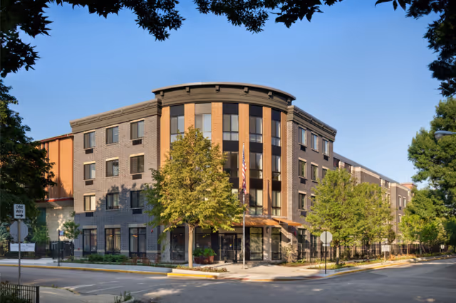 Exterior view of a modern four-story senior living facility building with brick and dark panel accents, surrounded by trees and located at a street corner with clear blue sky.