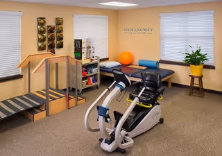 A small physical therapy or exercise room with a stationary recumbent bike in the foreground, a set of stairs with handrails on the left, a treatment table with a blue bolster and an orange exercise ball against the wall, and a plant on a wooden stand near a window. The walls are painted light yellow, and there is a motivational quote on the wall that reads 'Life is a journey, not a destination.'