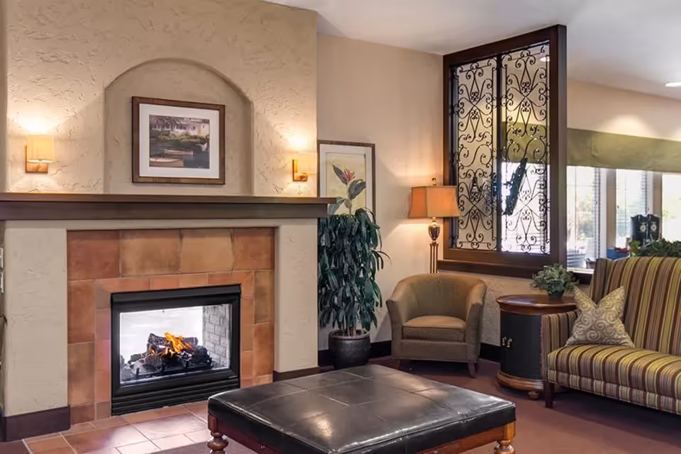 A cozy living room area with a lit fireplace surrounded by tan tiles and a textured beige wall. Above the fireplace is a framed picture, flanked by two wall sconces. To the right, there is a green armchair, a round wooden side table with a lamp, a potted plant, and a striped sofa with decorative pillows. A window with decorative wrought iron bars and a green valance is visible in the background.