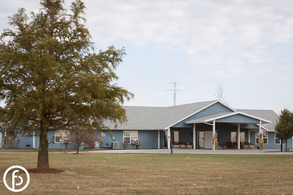 Exterior view of a single-story blue building with a gray roof, a covered entrance with chairs, and a large tree in the foreground on a grassy lawn under a partly cloudy sky.