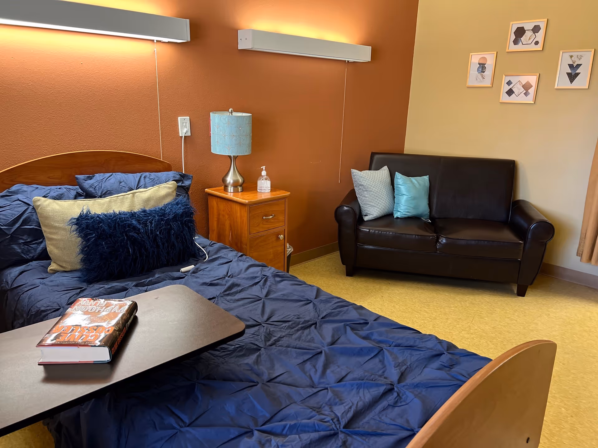 A furnished senior bedroom with a made bed covered in blue bedding, a wooden nightstand with a lamp and hand sanitizer, and a small black leather sofa against a brown wall.