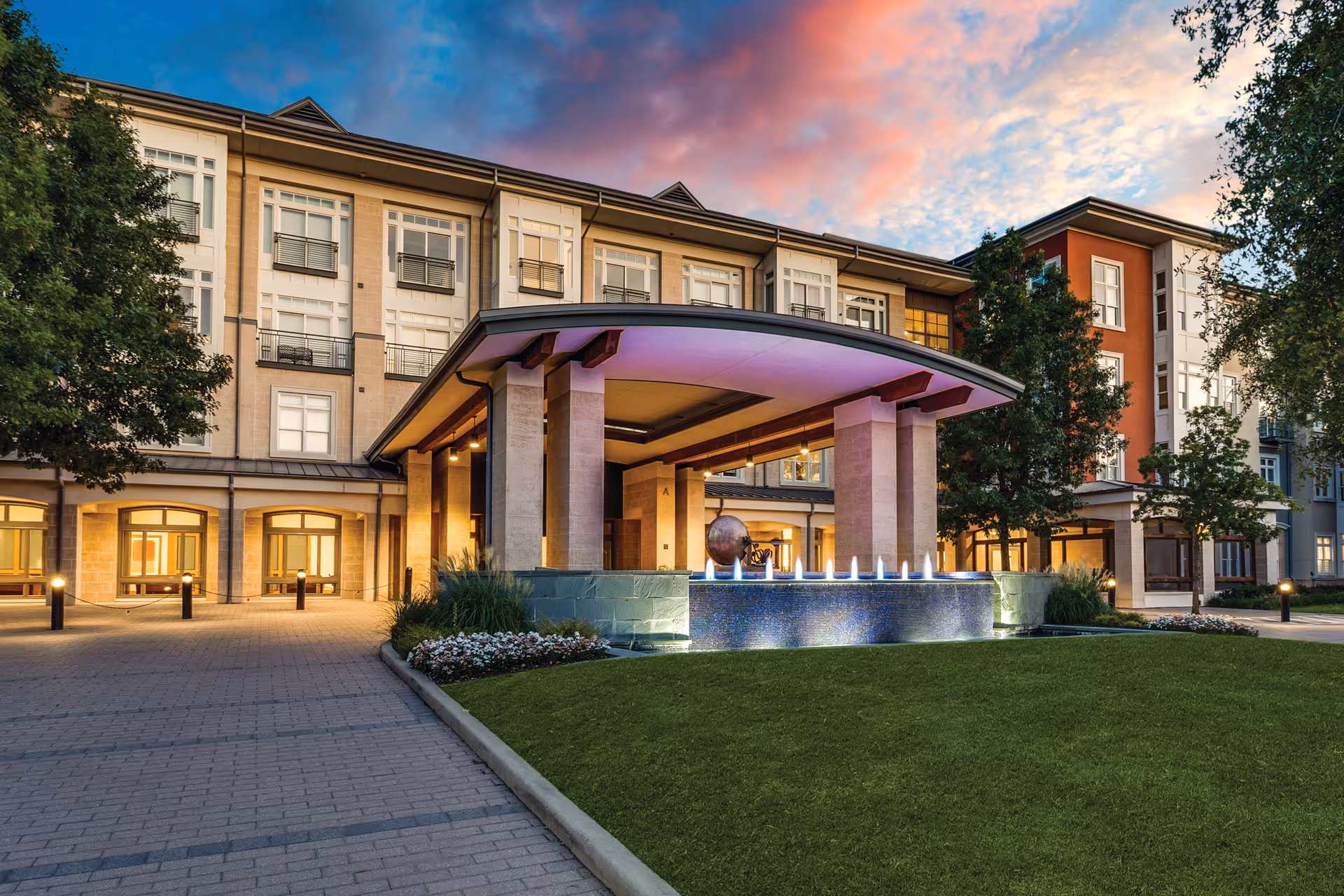 Exterior view of The Legacy Willow Bend senior living facility at dusk, featuring a modern entrance with a covered driveway, illuminated water fountain, well-maintained lawn, trees, and a colorful sky with clouds.
