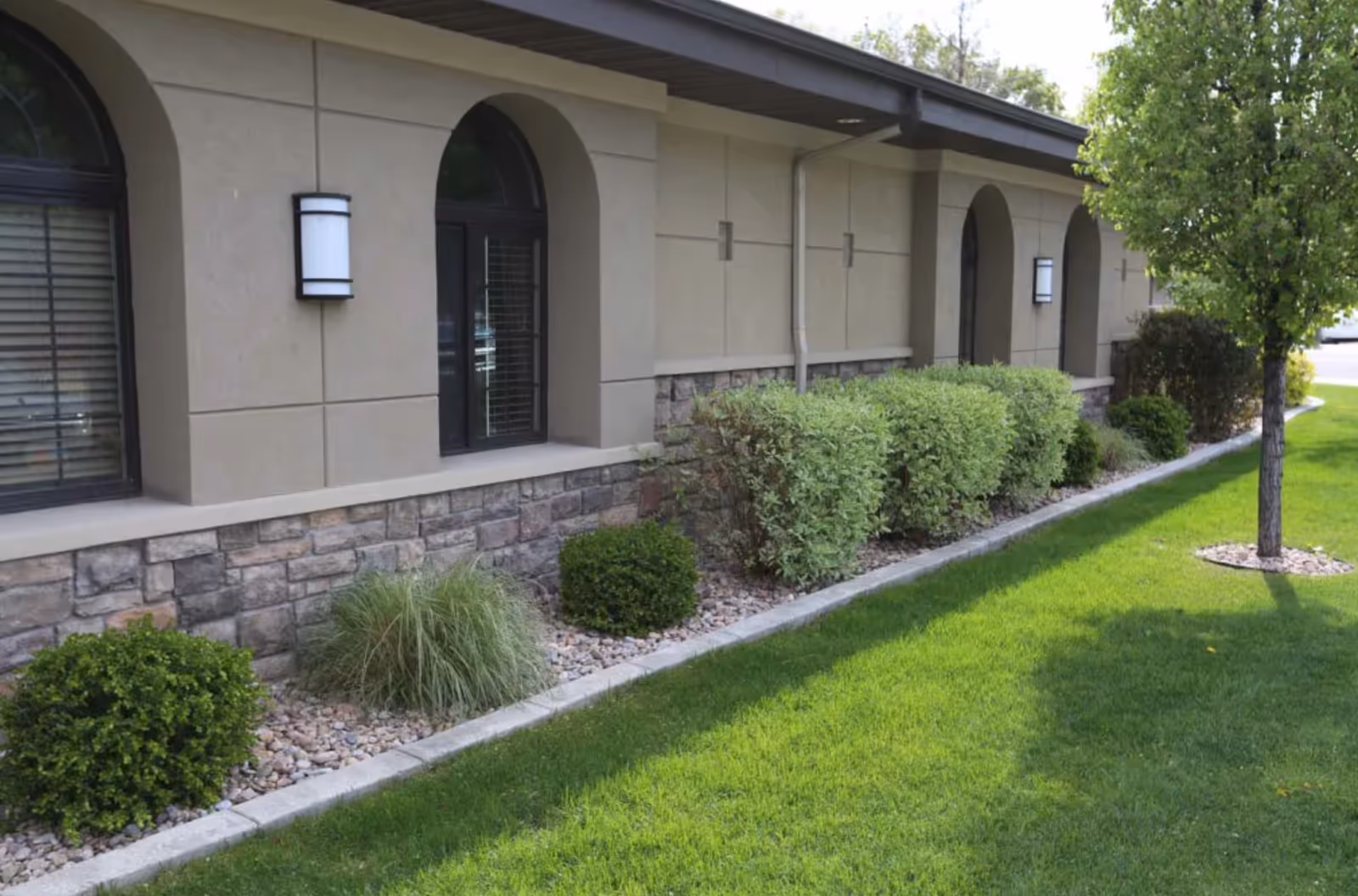 Exterior view of a building with beige walls, stone accents at the bottom, arched windows with black frames, wall-mounted lights, and a landscaped area with green bushes, grass, and a tree.