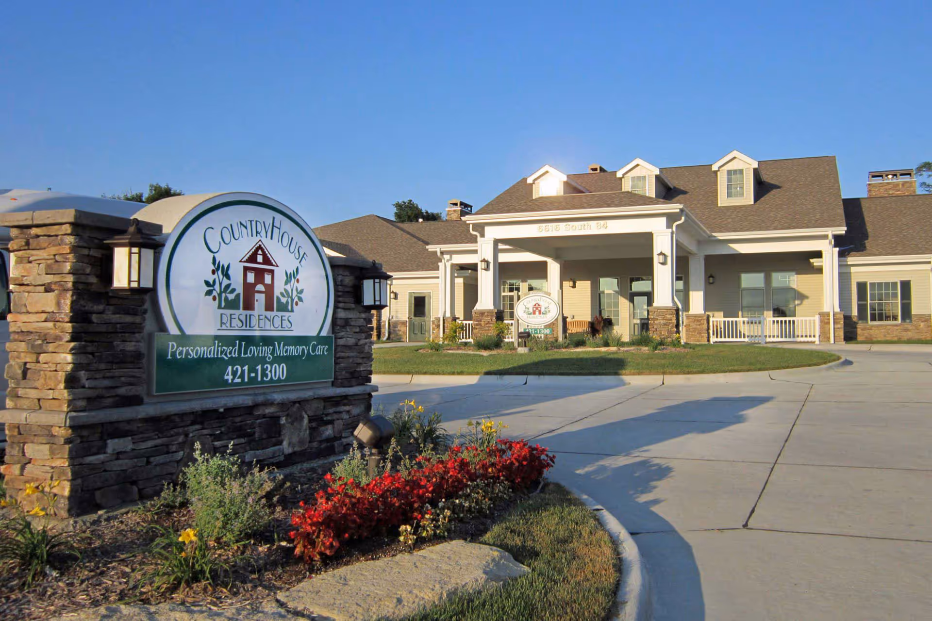 Exterior view of CountryHouse Residences, a senior living facility with a stone and white-paneled building, a covered entrance, and a large sign in front surrounded by flowers and landscaping.