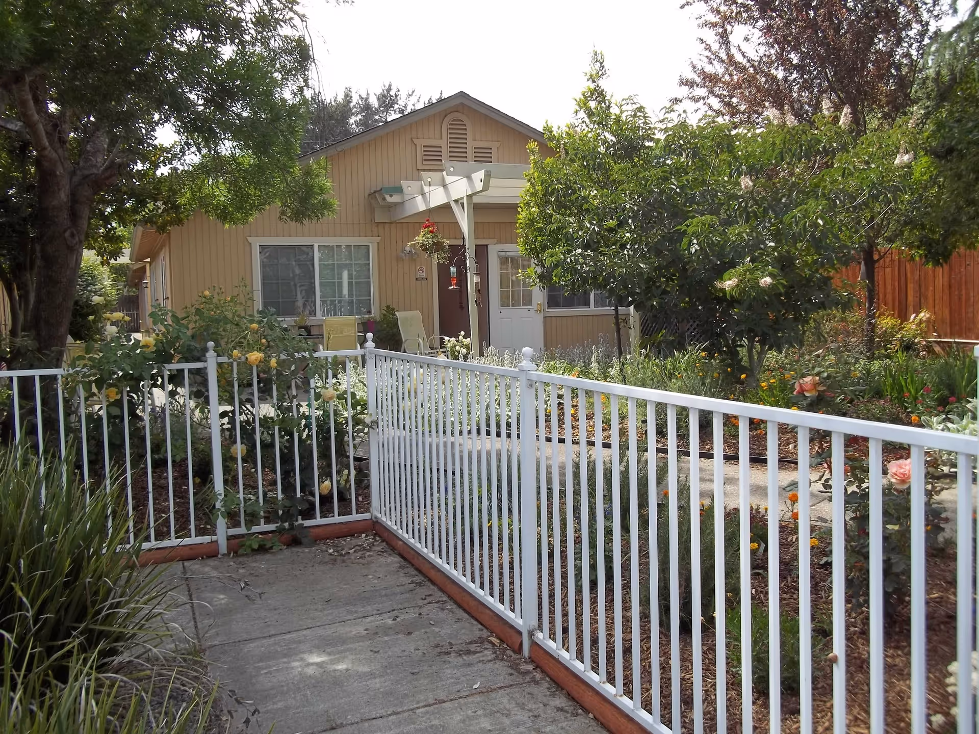 Front exterior of a single-story house with a white metal fence, garden beds and a pathway leading to the entrance.