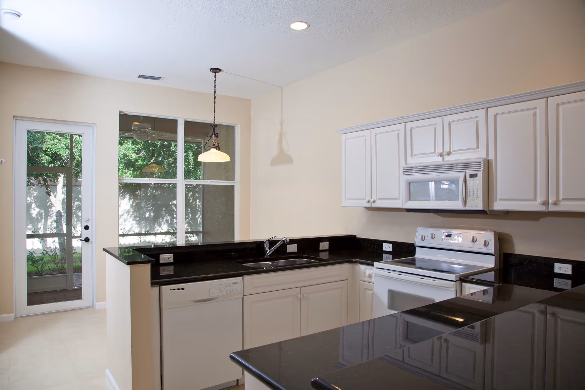 A modern kitchen with white cabinets, black granite countertops, a white electric stove with oven, a microwave above the stove, a dishwasher, and a sink. There is a glass door and large windows letting in natural light, with a hanging light fixture above the counter.