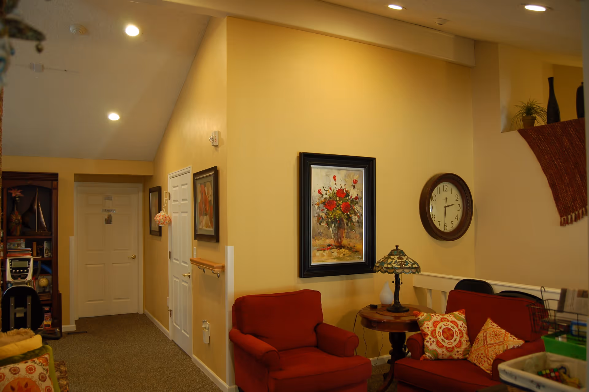 Interior of a cozy living room area with beige walls, a red armchair and a red loveseat adorned with patterned pillows. A round wooden side table with a decorative lamp sits between the chairs. A framed painting of flowers and a round wall clock are mounted on the wall. There is a hallway with closed white doors and framed artwork on the walls. The ceiling has recessed lighting.