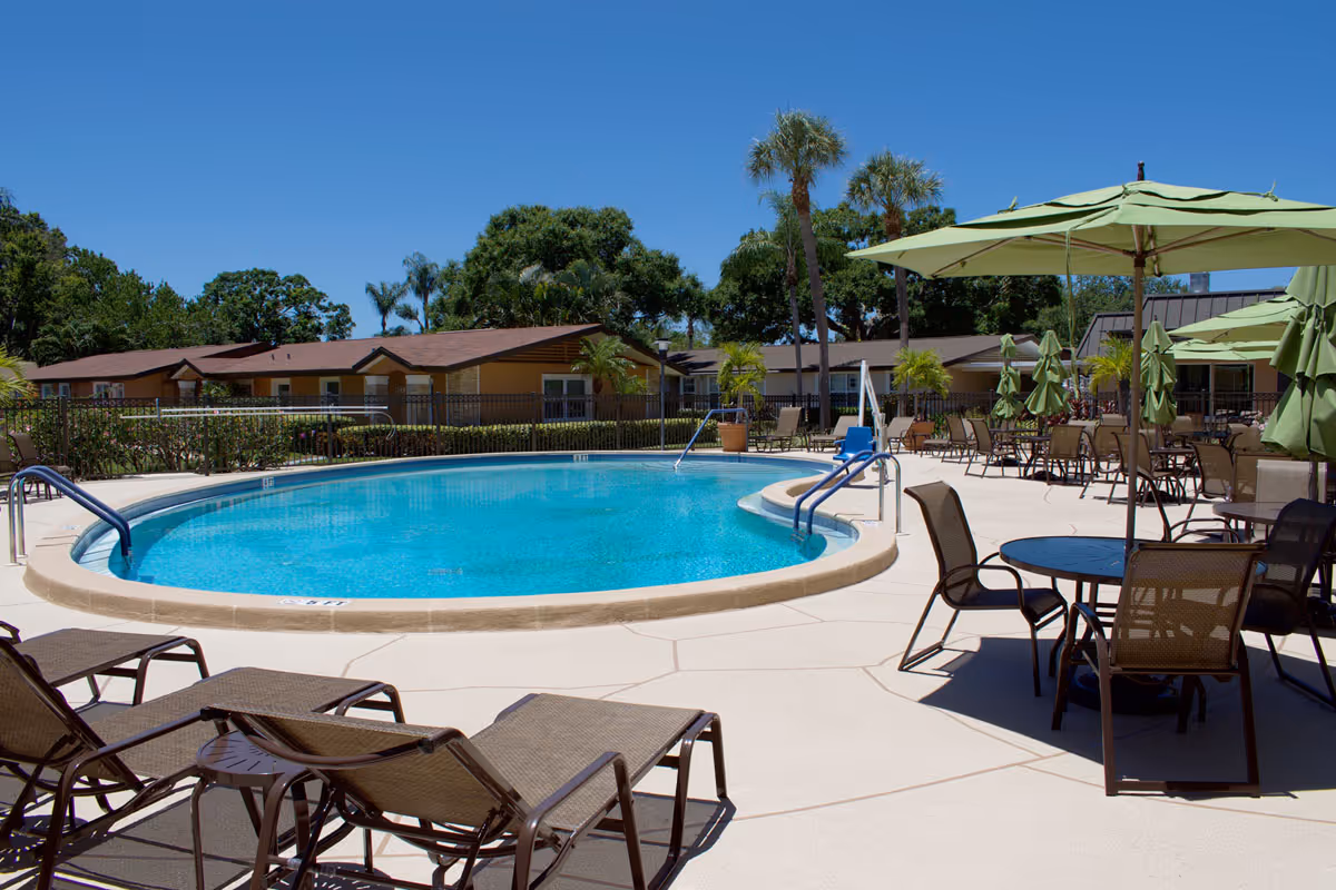 Outdoor swimming pool area with lounge chairs and tables with green umbrellas, surrounded by a fence and trees under a clear blue sky at Westminster Suncoast facility.
