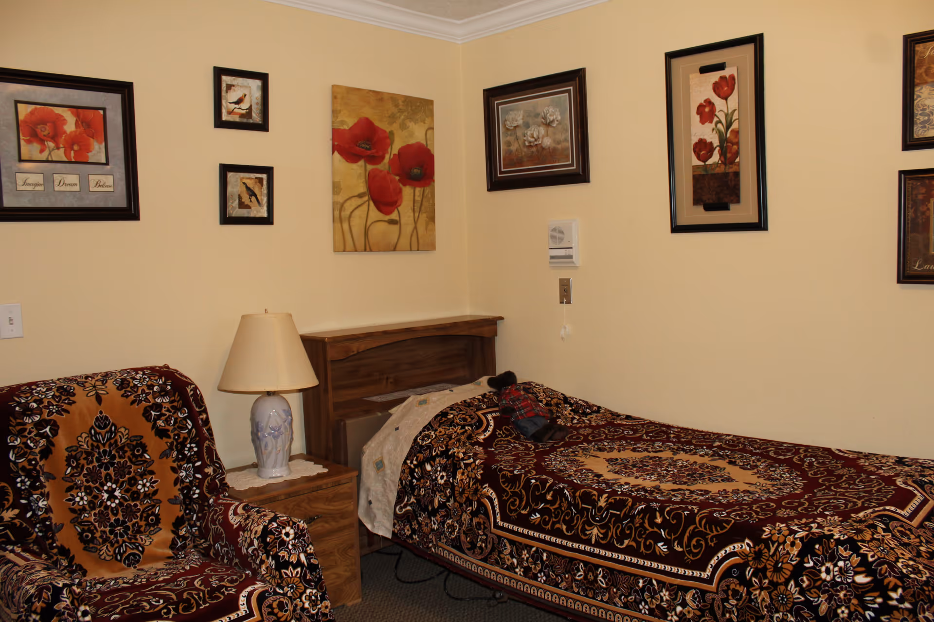 A cozy bedroom with a single bed covered in a patterned brown and beige bedspread. Next to the bed is a wooden nightstand with a white lamp. There is a matching armchair with a similar patterned cover. The walls are decorated with several framed floral and bird-themed paintings.