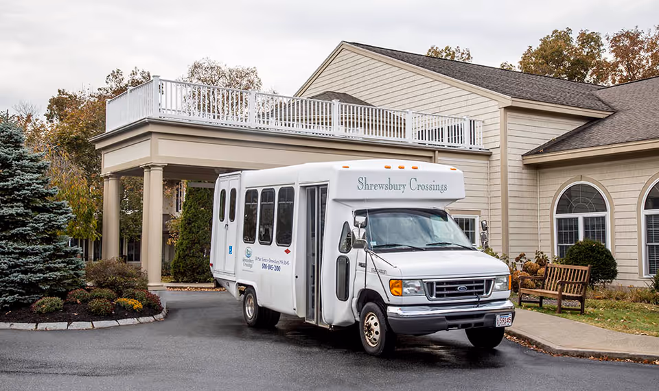 A white shuttle bus labeled 'Shrewsbury Crossings' parked in front of a beige senior living facility building with a covered entrance, landscaped bushes, and a wooden bench on the sidewalk.