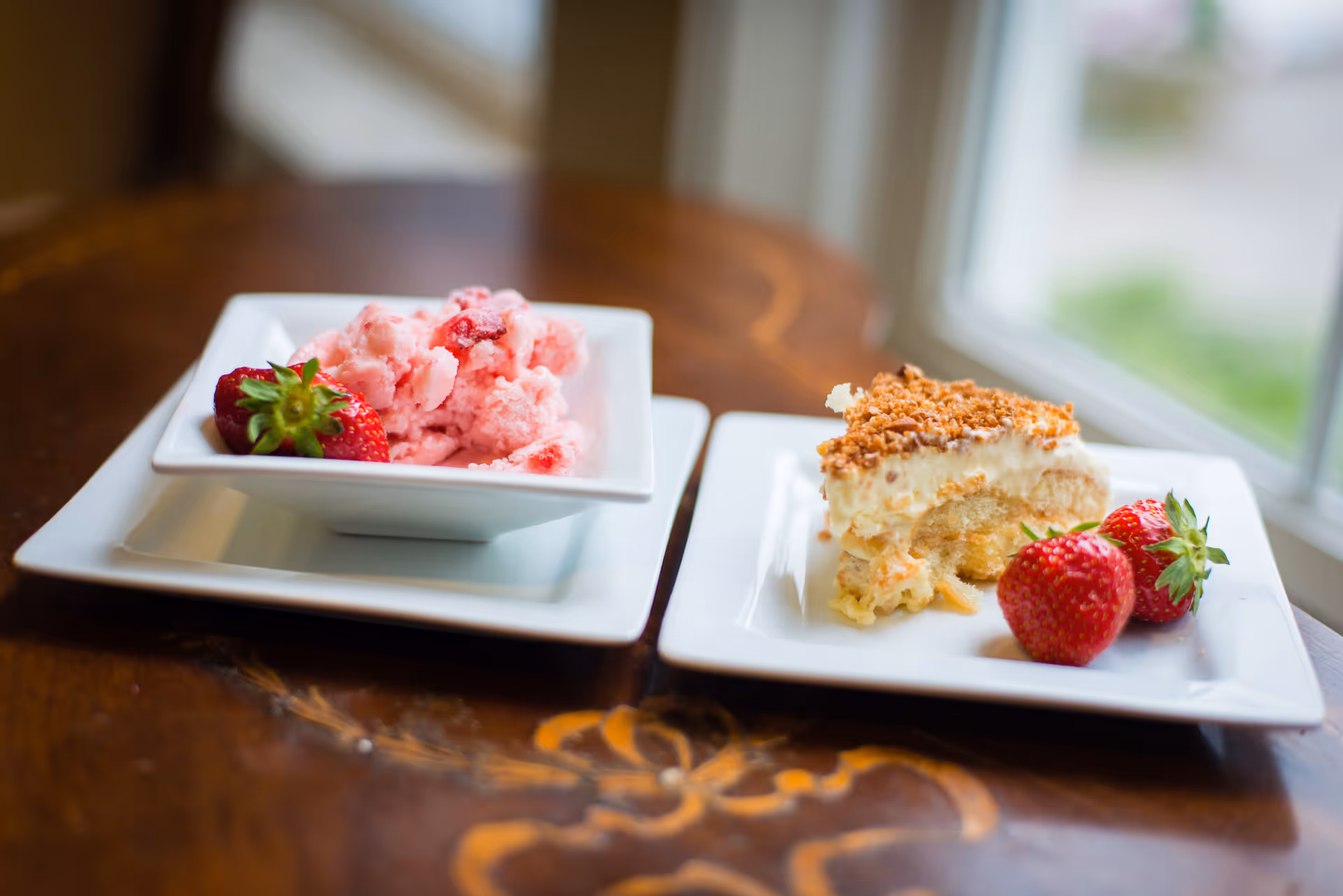 Two white square plates on a wooden table near a window. One plate holds a bowl of pink strawberry ice cream with a fresh strawberry on the side. The other plate has a slice of creamy dessert topped with crumbs and two fresh strawberries.