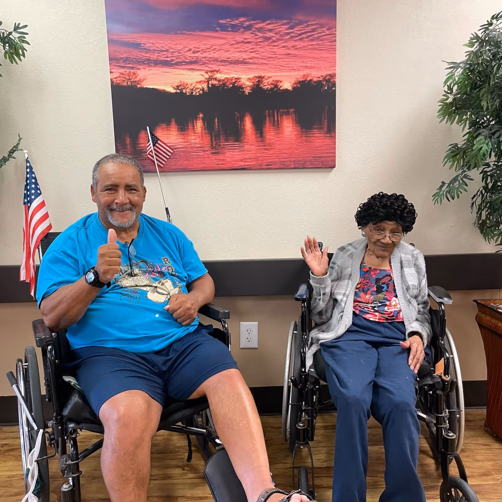 Two elderly individuals sitting in wheelchairs inside a room. The man on the left is wearing a blue t-shirt and shorts, giving a thumbs-up and smiling. The woman on the right is wearing glasses, a patterned shirt, and a gray cardigan, waving with her right hand. Behind them is a wall with a large framed picture of a sunset over a body of water and two small American flags attached to the wheelchairs. There are plants on either side of the room and a wooden floor.