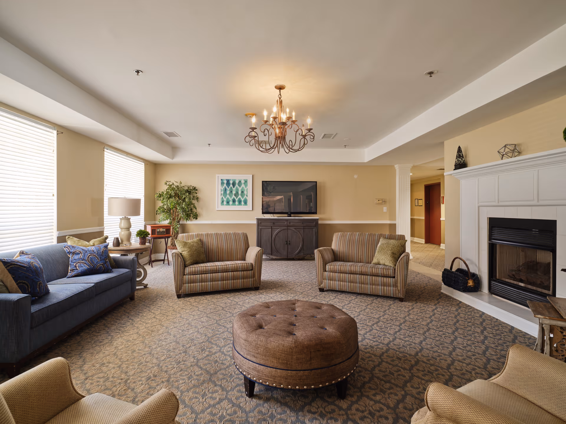 A cozy living room in an assisted living facility featuring a blue sofa with patterned pillows, two striped armchairs with green cushions, a round brown ottoman in the center, a fireplace on the right, a TV mounted on a cabinet against the far wall, a chandelier hanging from the ceiling, and large windows with blinds letting in natural light.