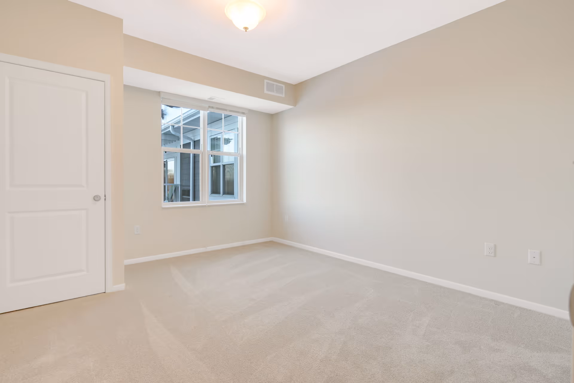 Empty room with beige carpet, beige walls, a white door on the left, and a window showing an exterior view of another building section.