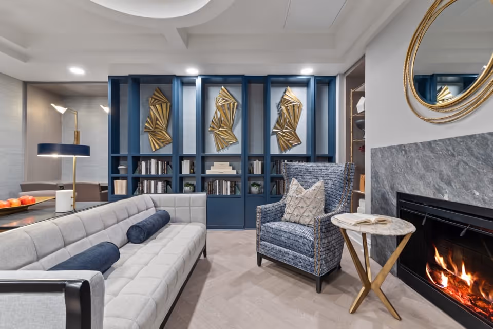 Stylish lounge with a light gray tufted sofa, patterned armchair, blue built-in shelving with gold wall art, and a lit fireplace.