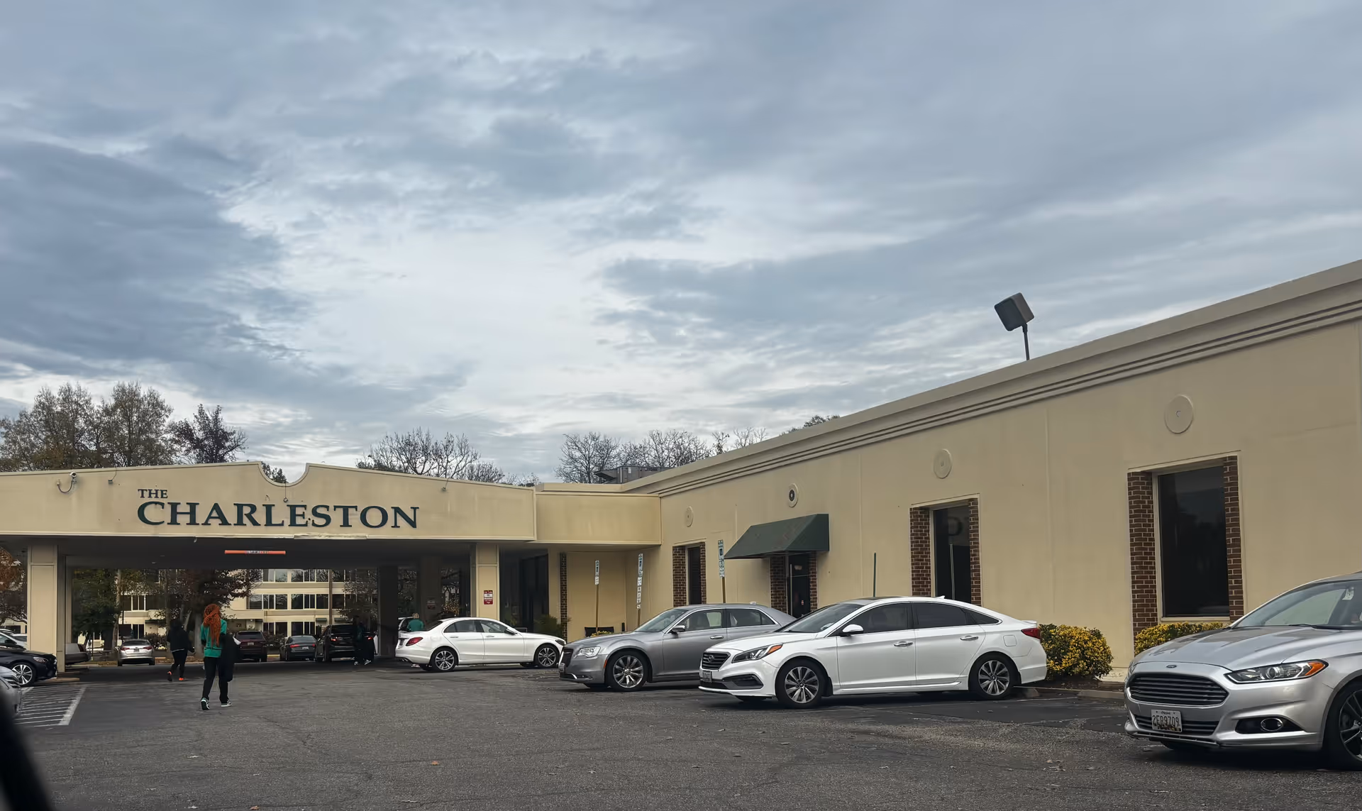Exterior view of The Charleston Senior Community building with a covered entrance. Several cars are parked in front of the building, and a few people are walking near the entrance. The sky is cloudy.