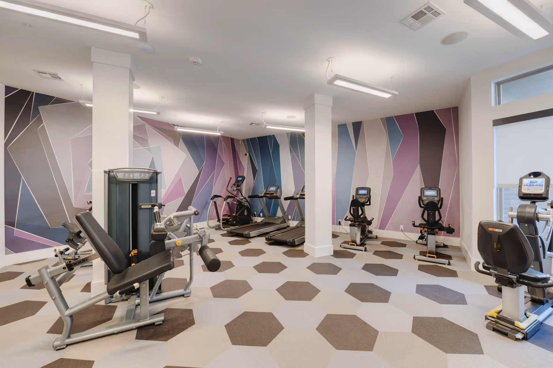 Bright fitness room with exercise machines, treadmills, and weight equipment against geometric patterned walls and hexagonal floor tiles.