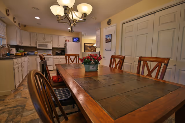 Interior view of a kitchen and dining area in an assisted living facility. The image shows a wooden dining table with a tiled surface and six wooden chairs around it. A small potted plant with red flowers is placed in the center of the table. The kitchen area in the background has white cabinets, a microwave, a refrigerator, and a stove. The floor has a stone tile pattern, and a chandelier with five lights hangs above the dining table.