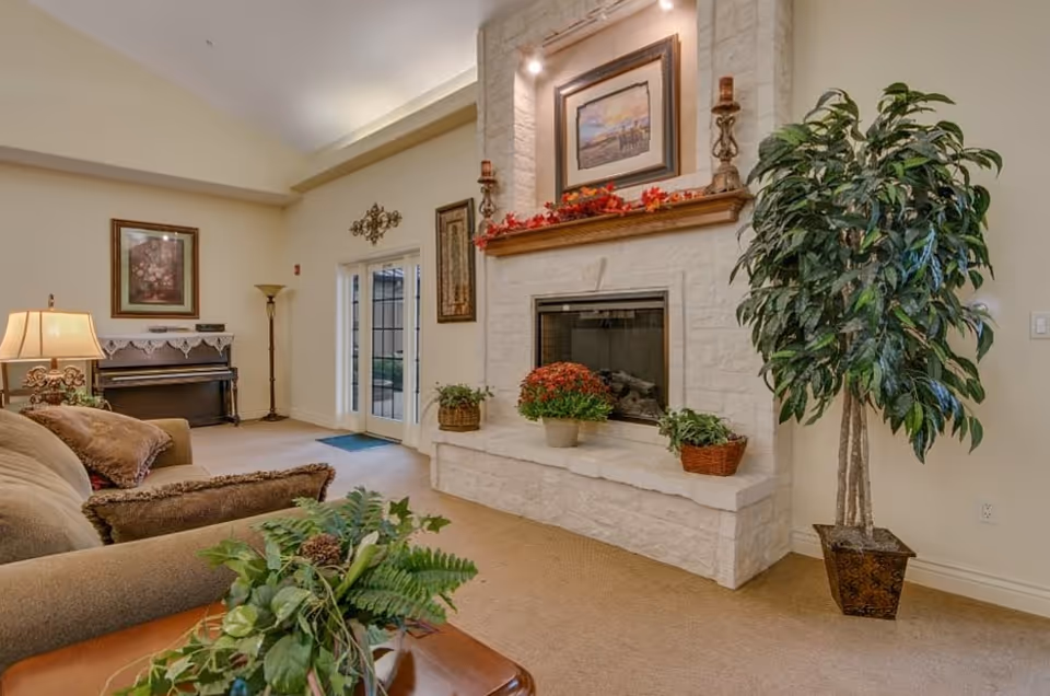 A cozy living room area with a beige sofa adorned with pillows, a large potted plant, and a white stone fireplace decorated with candles, framed artwork, and autumn-themed garland. There is a piano against the far wall with a framed picture above it, a floor lamp, and double glass doors leading outside.