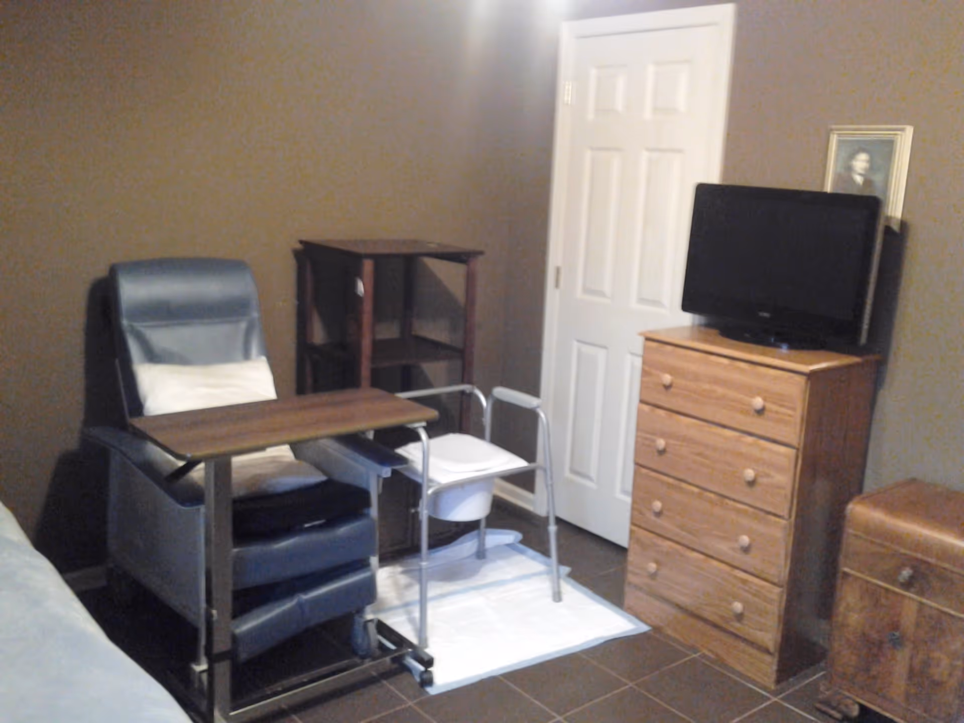 A room with brown walls and tiled floor featuring a blue recliner chair with a white pillow and an overbed table, a small wooden shelving unit, a white commode chair on a white mat, a wooden chest of drawers with a flat-screen TV on top, and a small wooden chest on wheels.
