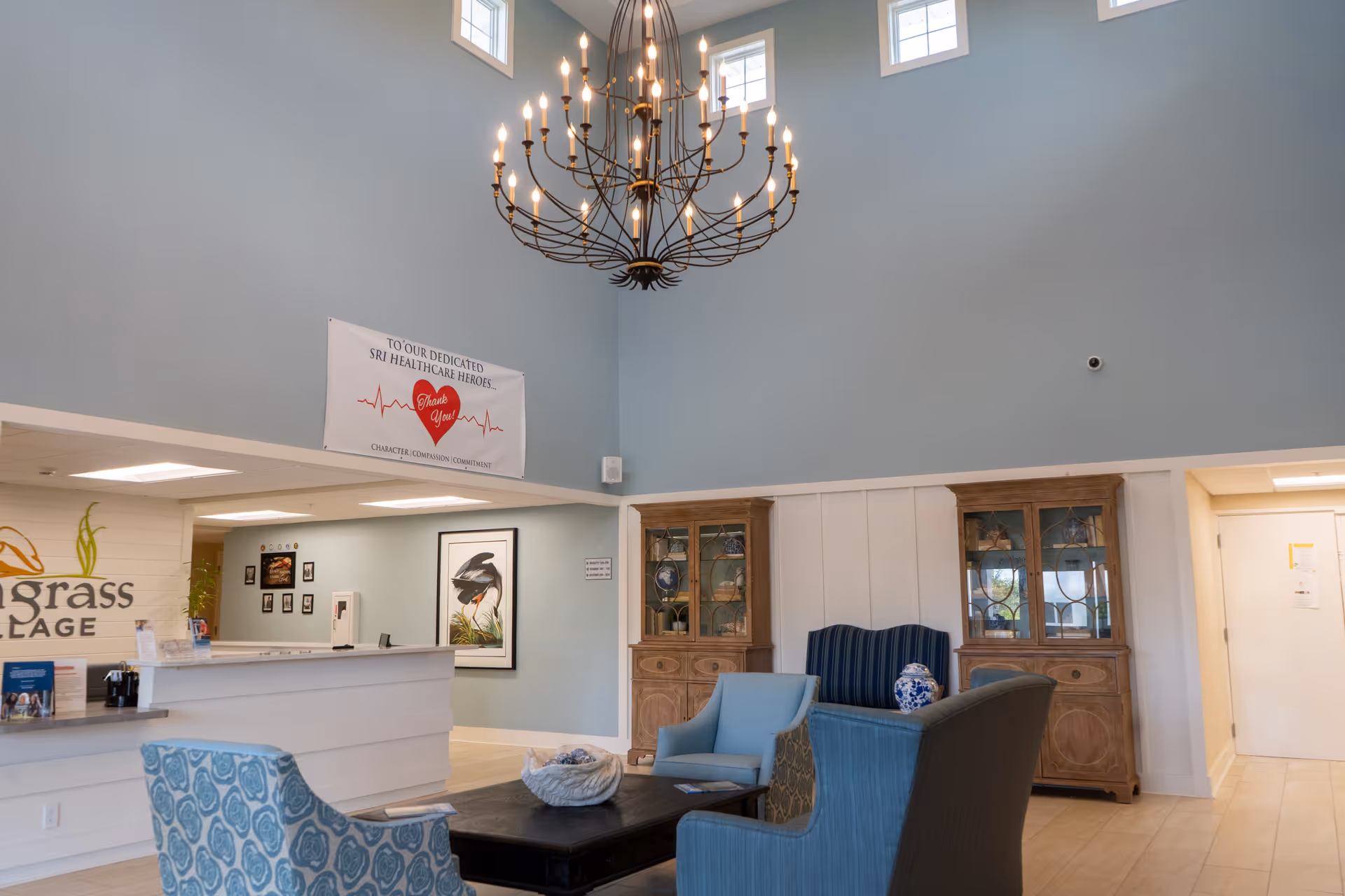 A senior living facility common area with high ceilings painted light blue, featuring a large chandelier. The room has comfortable seating including blue and patterned armchairs around a dark wooden coffee table. Two wooden display cabinets with glass doors are against the wall. A reception desk is visible to the left with a sign that partially reads 'grass VILLAGE'. A banner on the wall thanks healthcare heroes. The floor is light-colored tile.