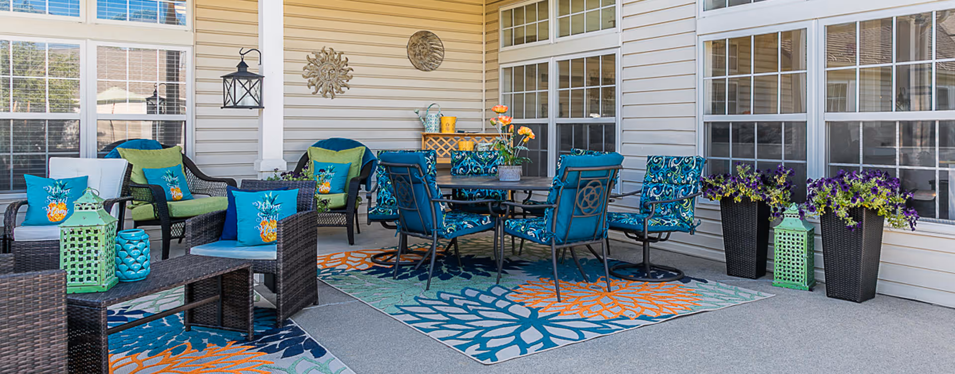 Outdoor patio area with wicker and metal chairs featuring blue and green cushions and pillows with pineapple designs, a round table, colorful floral rugs, decorative lanterns, potted flowers, and wall-mounted metal sun decorations on the beige siding of the building.