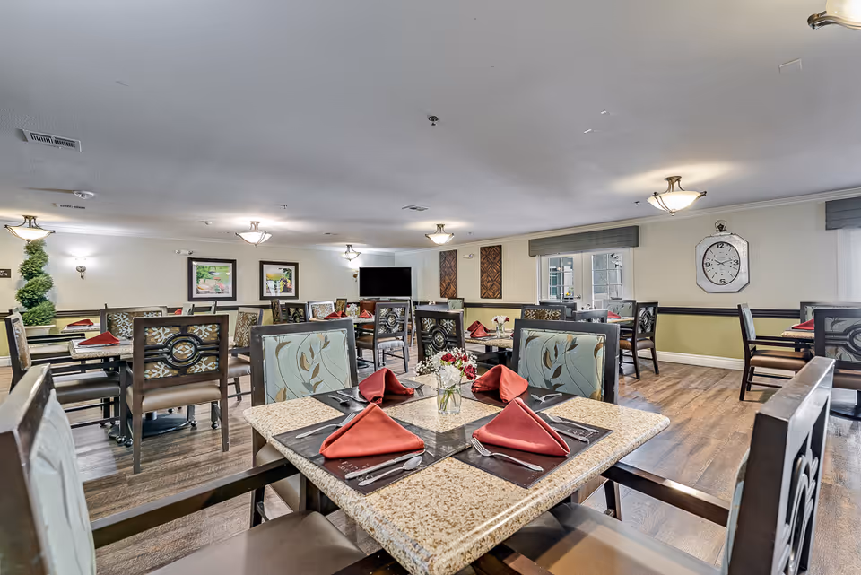 Communal dining room with multiple set tables and upholstered chairs, red napkins, a wall clock and a television.