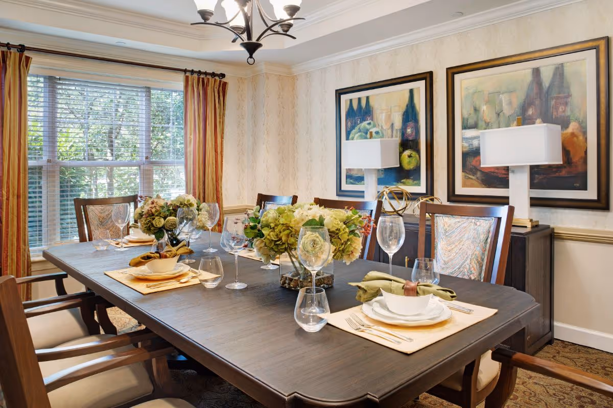 A well-lit dining room with a large wooden table set for six people. The table is decorated with floral centerpieces, wine glasses, plates, and napkins. The room features a large window with striped curtains, two framed paintings on the wall, and a sideboard with two table lamps.