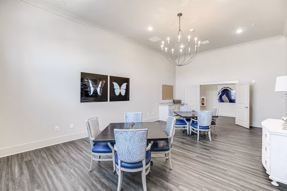 A bright, modern dining room with two square tables, each surrounded by four blue cushioned chairs. The room features light gray walls, wood-look flooring, a contemporary chandelier, and butterfly artwork on the wall. There is a small counter area with a basket and an open doorway leading to another room with artwork visible inside.