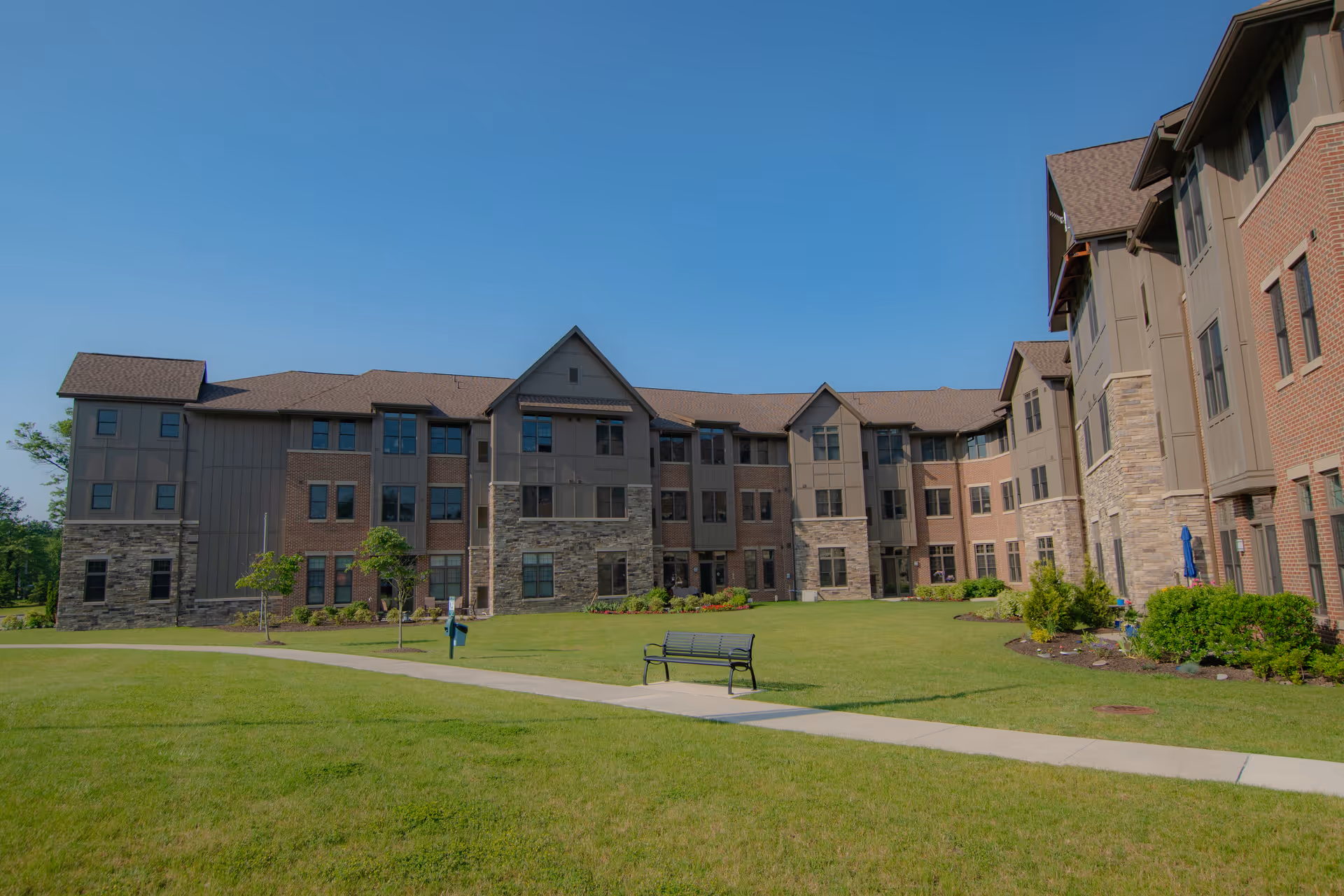 Exterior view of a multi-story senior living facility building with a mix of brick and stone facade, surrounded by a well-maintained lawn and a paved walkway with a bench. The sky is clear and blue.