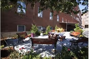 Outdoor patio area with several wooden benches and a round table with chairs, surrounded by plants and flowers, next to a brick building.