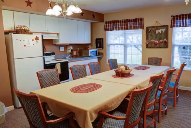 Communal dining area with a long table and multiple chairs, placemats, and a small kitchenette against the back wall.