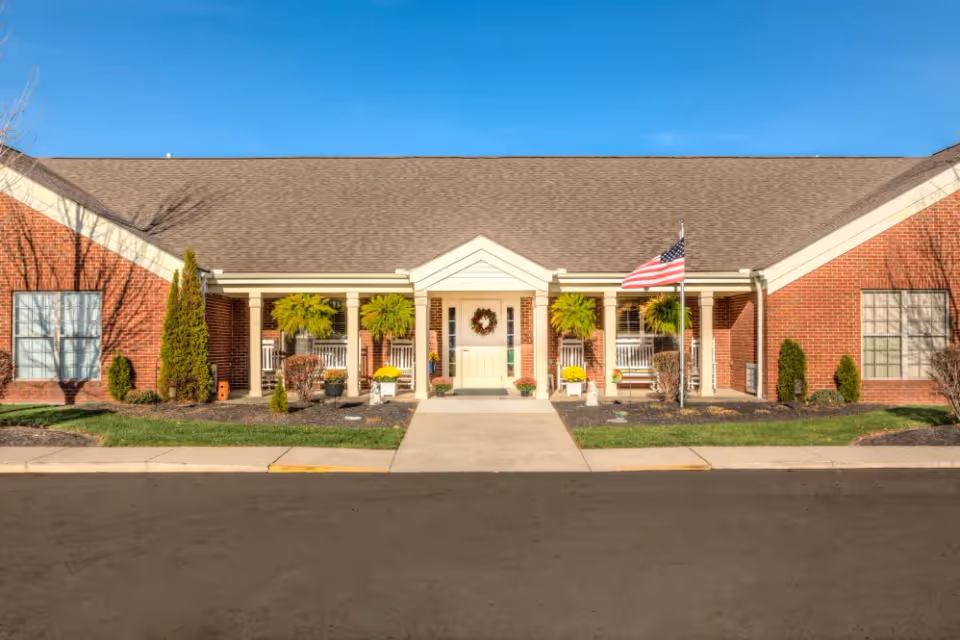 Front entrance of a single-story brick senior living building with a covered porch, potted plants, and an American flag.