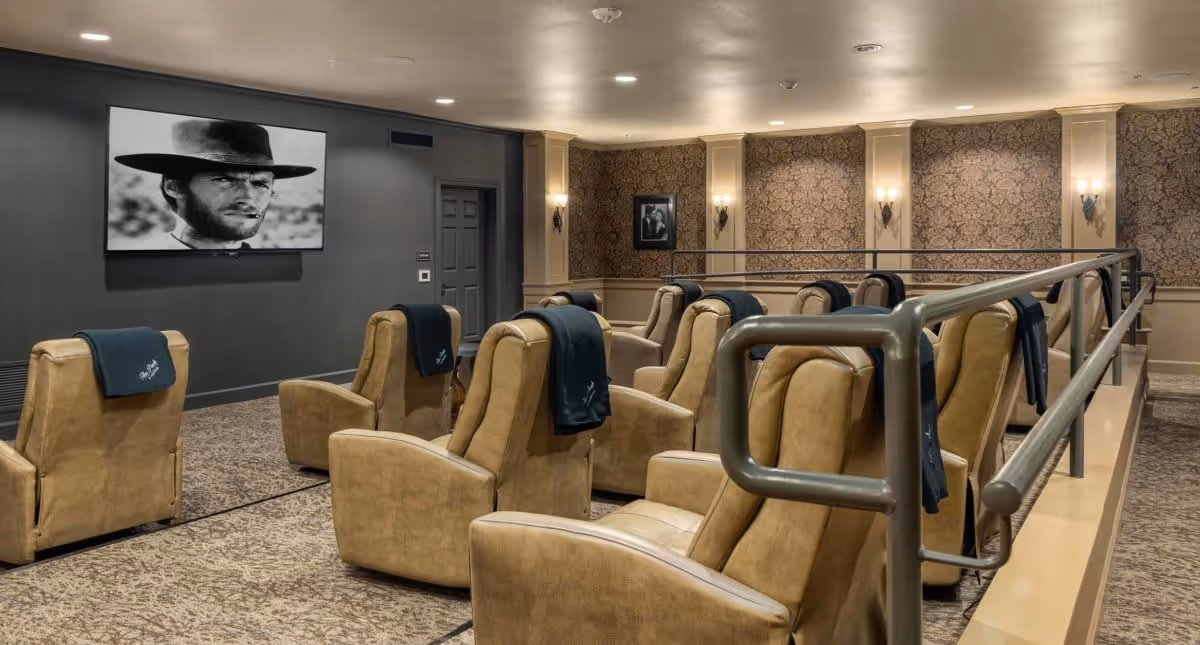 A cozy movie theater room with multiple tan leather recliner chairs arranged in rows facing a large screen displaying a black and white image of a man wearing a hat. The room has patterned wallpaper, wall sconces providing soft lighting, and a metal railing along the right side.