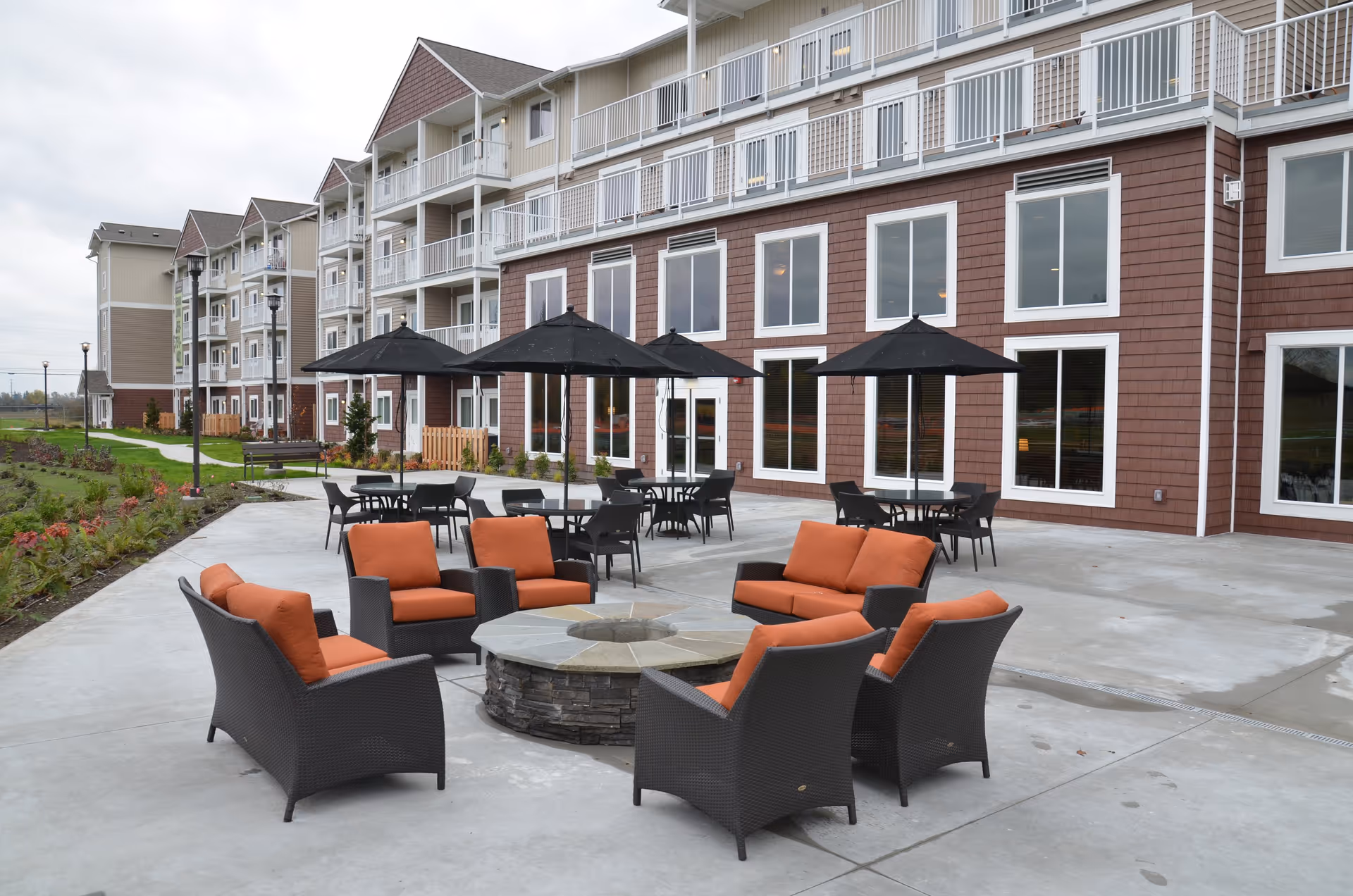 Outdoor patio area at Vintage at Lakewood featuring a circular stone fire pit surrounded by six wicker chairs with orange cushions. Behind the fire pit are several tables with black umbrellas and chairs. The patio is adjacent to a multi-story building with large windows and balconies.