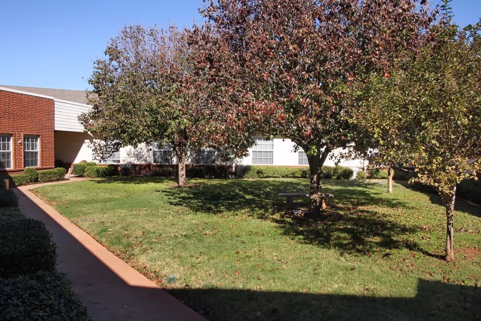 Outdoor courtyard area with green grass, several trees with some autumn-colored leaves, a concrete walkway, and a brick and white building in the background under a clear blue sky.
