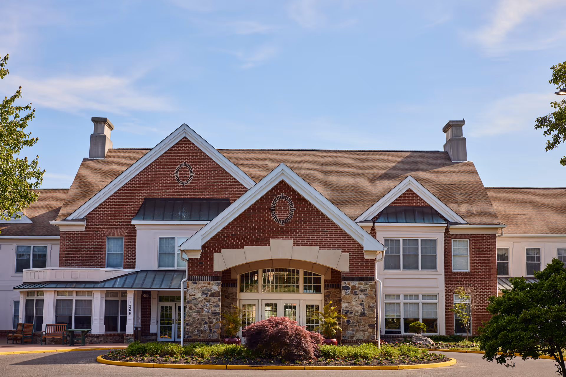 Front exterior view of a senior living facility building with red brick and stone accents, large windows, and a peaked roof under a blue sky with some clouds. There are landscaped plants and trees around the entrance area.