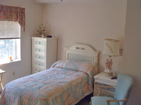 A small bedroom with a single bed covered in a pastel-colored quilt. There is a white headboard, a white chest of drawers in the corner, a nightstand with a decorative lamp and figurine, and a light blue upholstered chair. A window with blinds and floral valance lets in natural light.