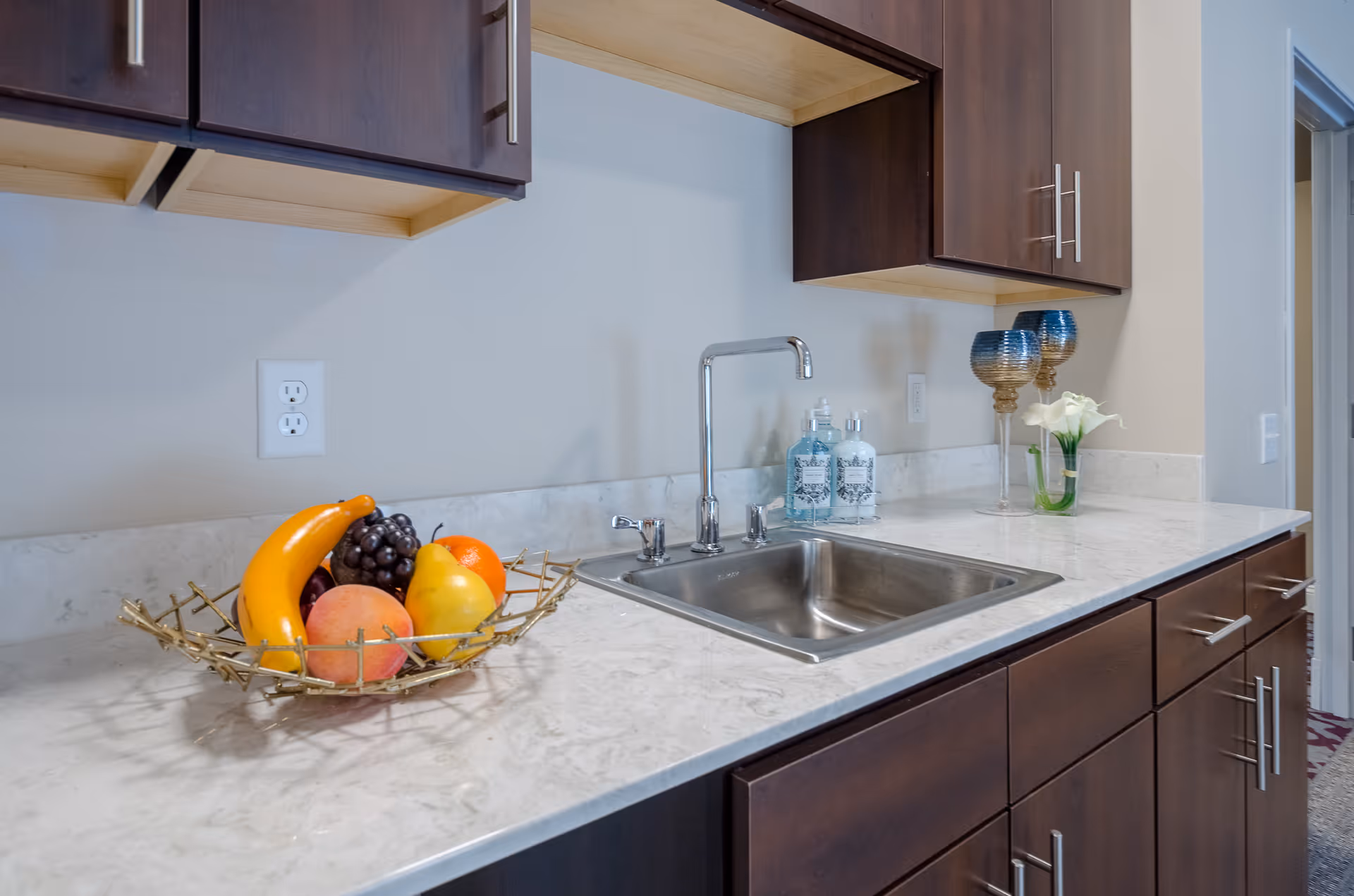 A kitchen countertop with a stainless steel sink and faucet. On the left side of the counter, there is a decorative fruit basket containing bananas, grapes, an orange, a pear, and a peach. On the right side, there are two decorative blue and gold goblets and a small vase with white flowers. Above the counter are dark wooden cabinets with silver handles.