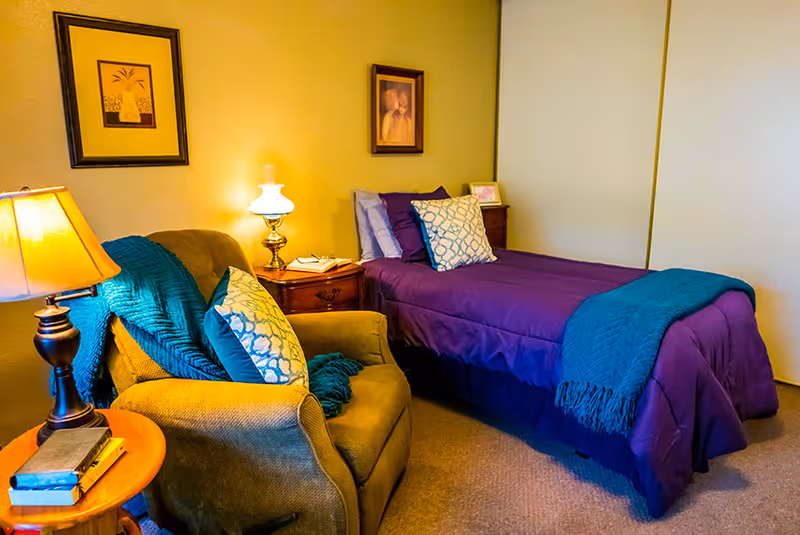 A cozy bedroom with a single bed dressed in purple bedding and a teal throw blanket. Next to the bed is a wooden nightstand with a lamp and some books. There is a comfortable armchair with teal cushions and a throw blanket. The walls are decorated with two framed pictures.