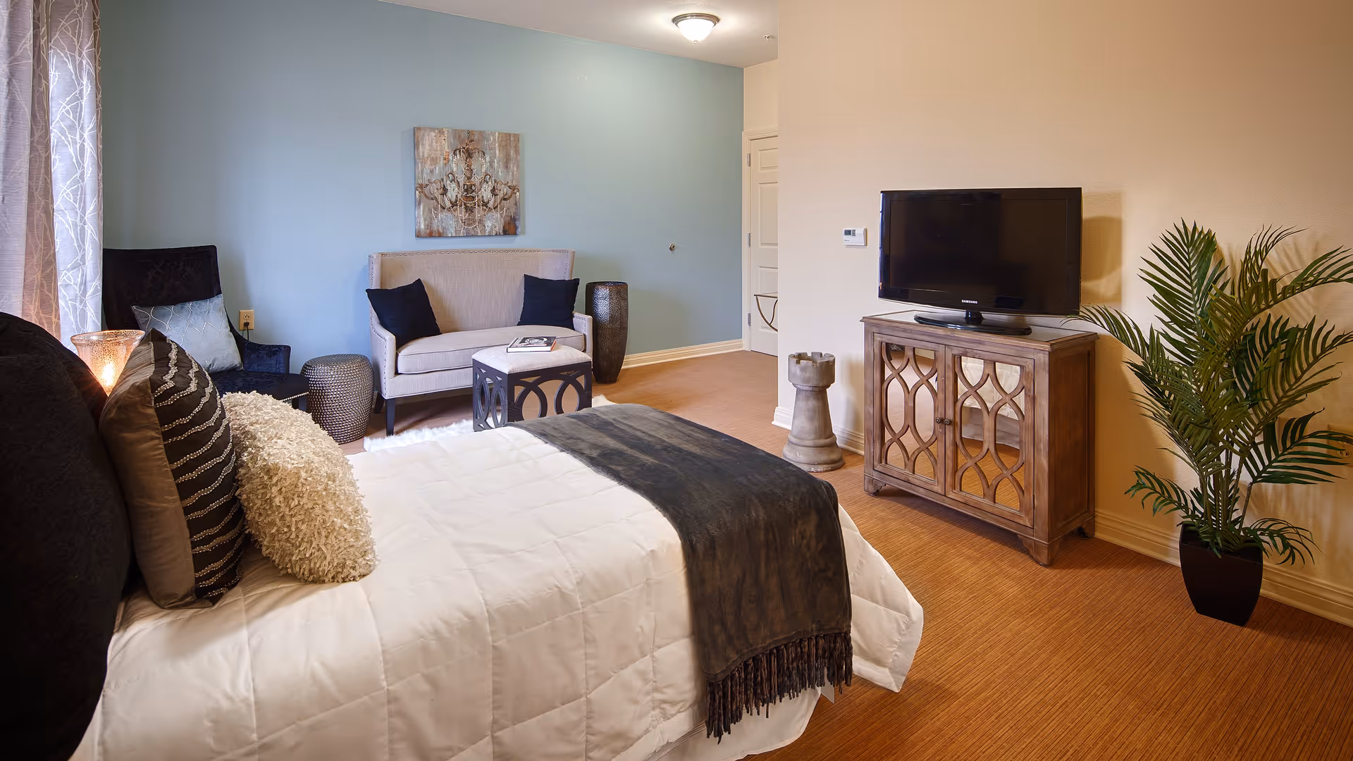 A cozy bedroom with a neatly made bed featuring multiple pillows and a dark throw blanket. In the background, there is a small sitting area with a loveseat, two black pillows, a side table, and a decorative wall art. A wooden cabinet with a flat-screen TV on top is positioned against the wall, next to a potted plant. The room has warm lighting and a soft blue accent wall.