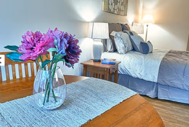 A cozy bedroom with a neatly made bed featuring multiple pillows and a gray comforter. Next to the bed is a wooden nightstand with a lamp and a book. In the foreground, there is a wooden table with a light blue woven runner and a glass vase holding vibrant purple and pink flowers.