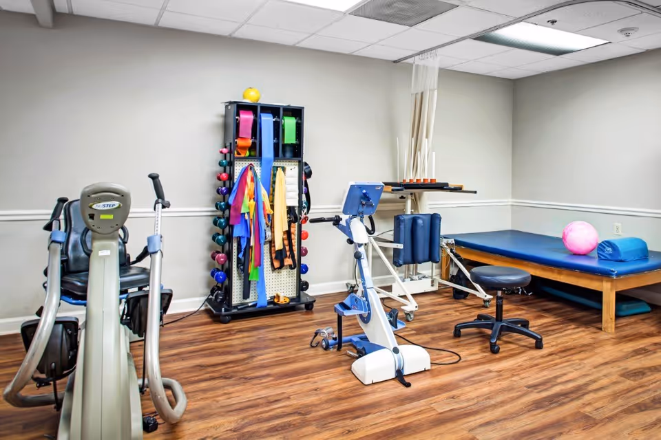 A physical therapy or exercise room with various rehabilitation equipment including a stationary bike, resistance bands, dumbbells, a padded treatment table with a pink exercise ball, and a stool. The room has wood flooring, white walls, and a drop ceiling with fluorescent lighting.