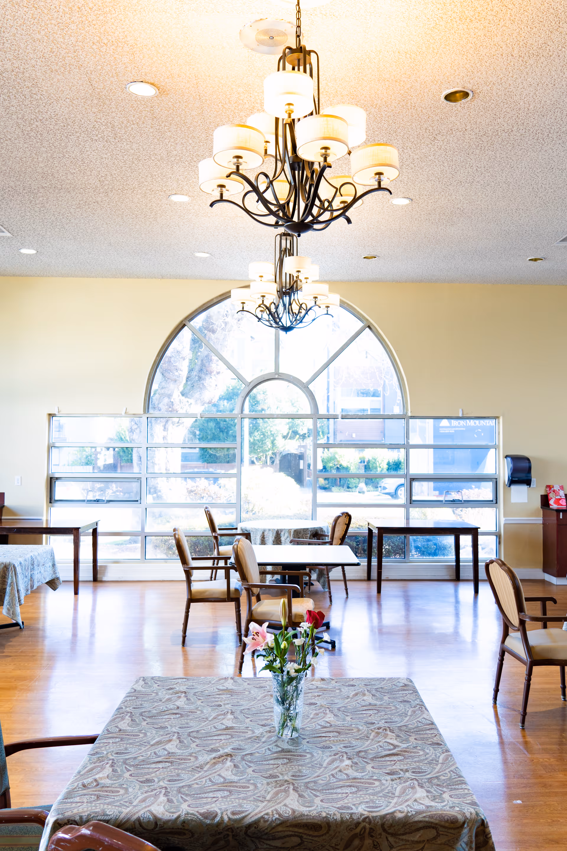 A bright dining room with several tables covered with patterned tablecloths and chairs arranged around them. A vase with flowers is placed on the nearest table. Large arched windows allow natural light to fill the room, and two elegant chandeliers hang from the ceiling.