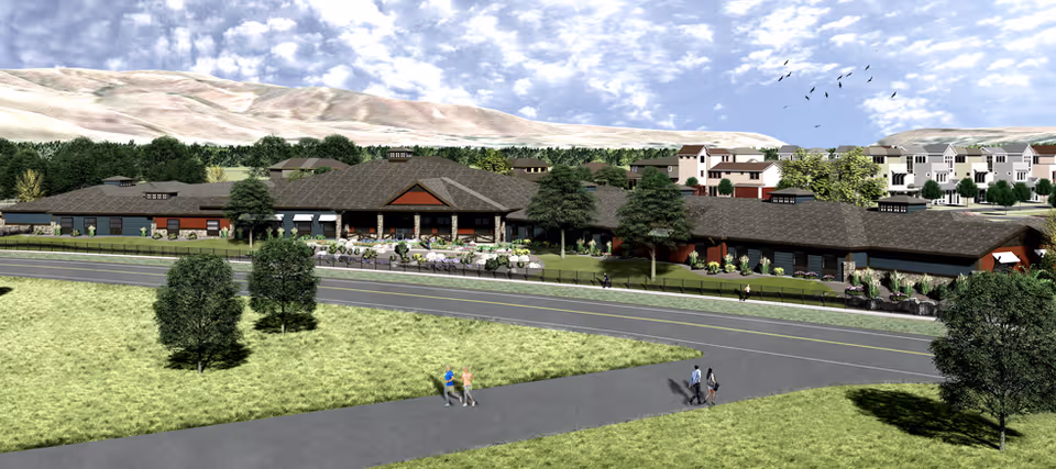 Exterior view of a large senior living facility with a dark roof and stone accents, surrounded by green lawns, trees, and a paved road. Several people are walking on the sidewalks near the building. In the background, there are hills and additional residential buildings under a partly cloudy sky.